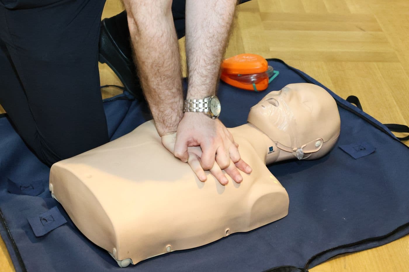 Hands performing chest compressions on a training manikin beside an AED